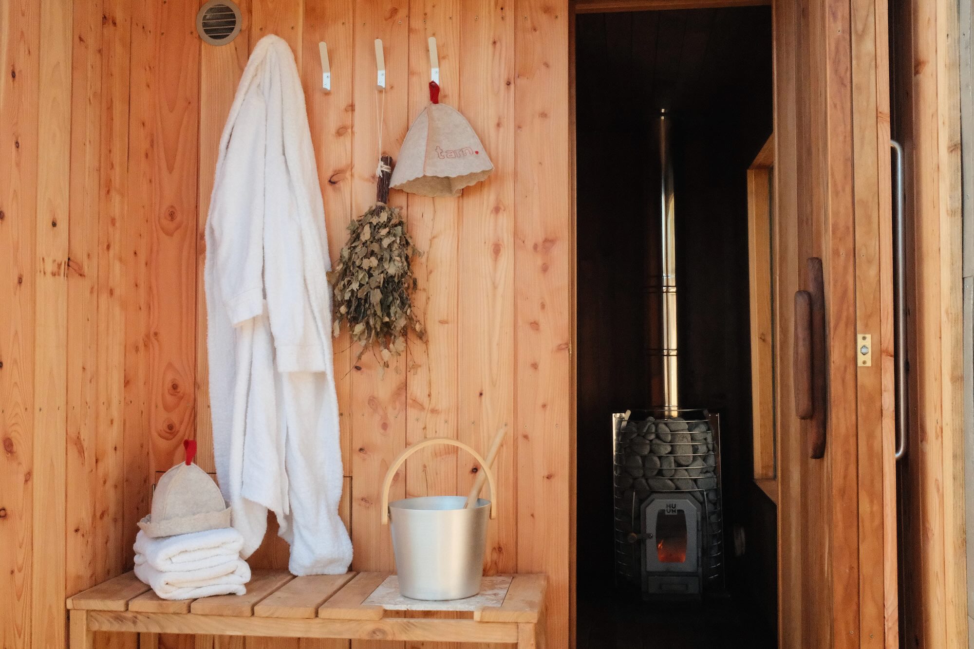 Wood-fired sauna interior with warm wooden walls, folded towels, silver bucket for ice and a fluffy white robe beside the entrance to the heated sauna room. A traditional sauna stove filled with wood glows inside, creating a calm and welcoming lakeside wellbeing setting.
