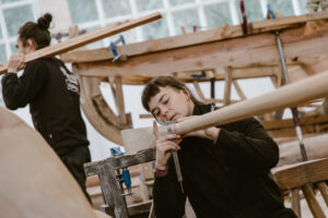 A person working on a part of a wooden boat in a workshop with someone else walking away behind them