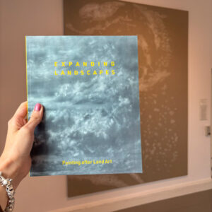 Hand holding Expanding Landscapes exhibition catalogue inside the exhibition gallery.