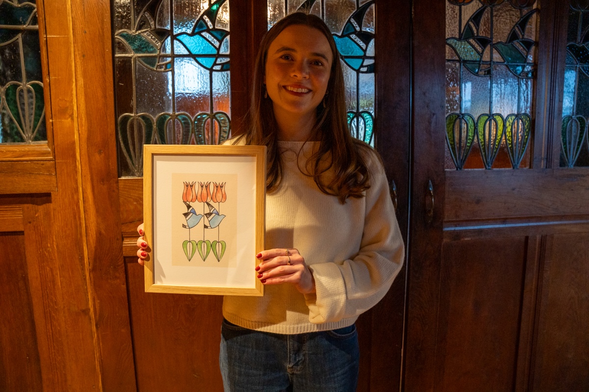 Artist happily holding their framed print of their contemporary stylised version of the tulip and blue bird stained glass windows featured in the background where they took the photo.