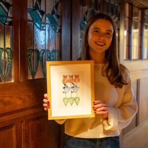 Artist Tiana McInerney holding a framed illustration of stylised tulip and blue bird design inspired by Blackwell’s stained glass window in the corridor, standing beside the decorative glass panels inside the Arts & Crafts house.