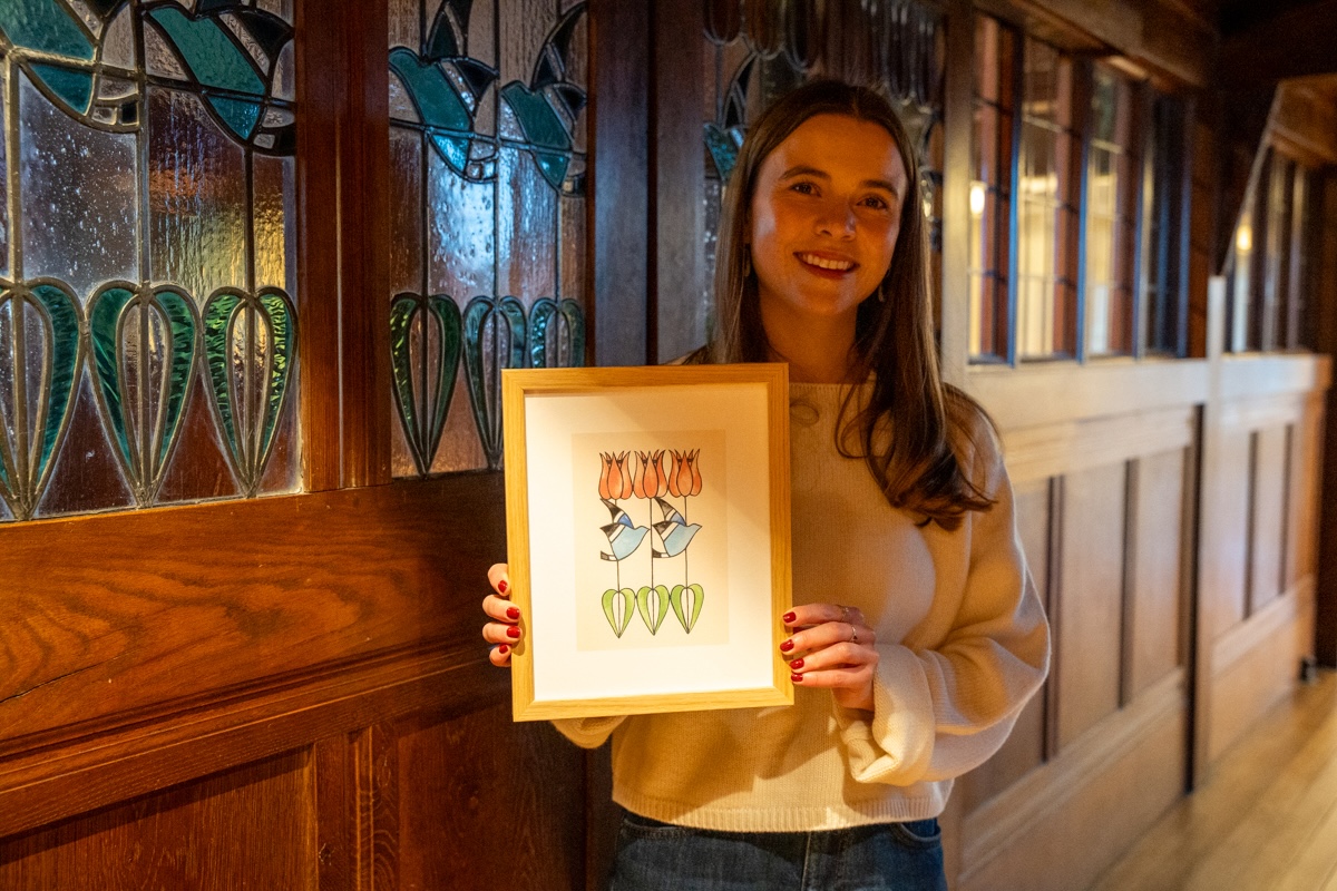 Tiana McInerney at Blackwell lo-res Credit Natasha Suriya from Lakeland Arts-036. Artist Tiana McInerney holding a framed illustration of stylised tulip and blue bird design inspired by Blackwell’s stained glass window in the corridor, standing beside the decorative glass panels inside the Arts & Crafts house.