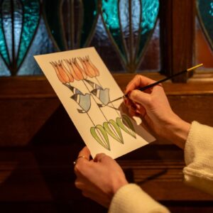 Artist Tiana McInerney painting a stylised tulip and leaf motif on card in front of the stained glass windows at Blackwell – the Arts & Crafts house.