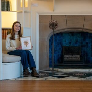 Artist Tiana McInerney seated beside the detailed inglenook fireplace in the White Drawing Room at Blackwell house, holding a framed illustration inspired by the iron and enamelled fire dogs on display.