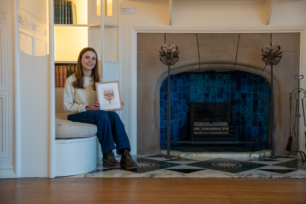 Tiana McInerney at Blackwell lo-res Credit Natasha Suriya from Lakeland Arts-098. Artist Tiana McInerney seated beside the detailed inglenook fireplace in the White Drawing Room at Blackwell house, holding a framed illustration inspired by the iron and enamelled fire dogs on display.