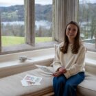 Artist Tiana McInerney sitting by the bay window at Blackwell – the Arts & Crafts house, holding one of her illustrated designs inspired by the building’s decorative motifs, with Windermere visible outside.