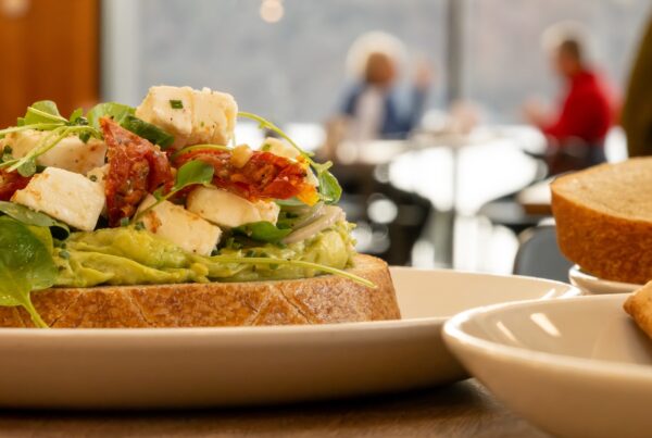 Zoomed in shot of creamy avocado, topped with red sundried cherry tomatoes, refreshing feta cheese, and garnished with leaves on a toasted white farmhouse slice of bread. In the distance you can see a couple enjoying a cup of coffee together.