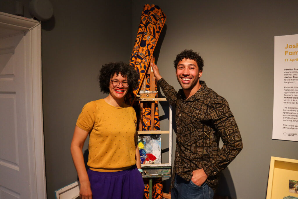Two artists smiling beside a painted easel in a gallery space, with exhibition text on the wall behind them.