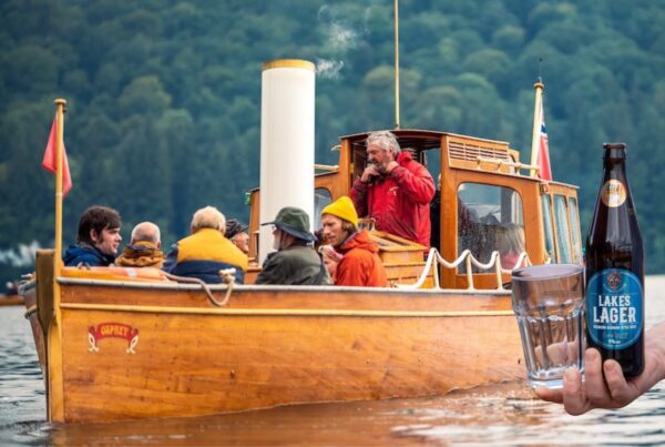 Osprey filled with happy passengers sailing across Windermere with a cutout of a hand holding a local craft beer and glass.